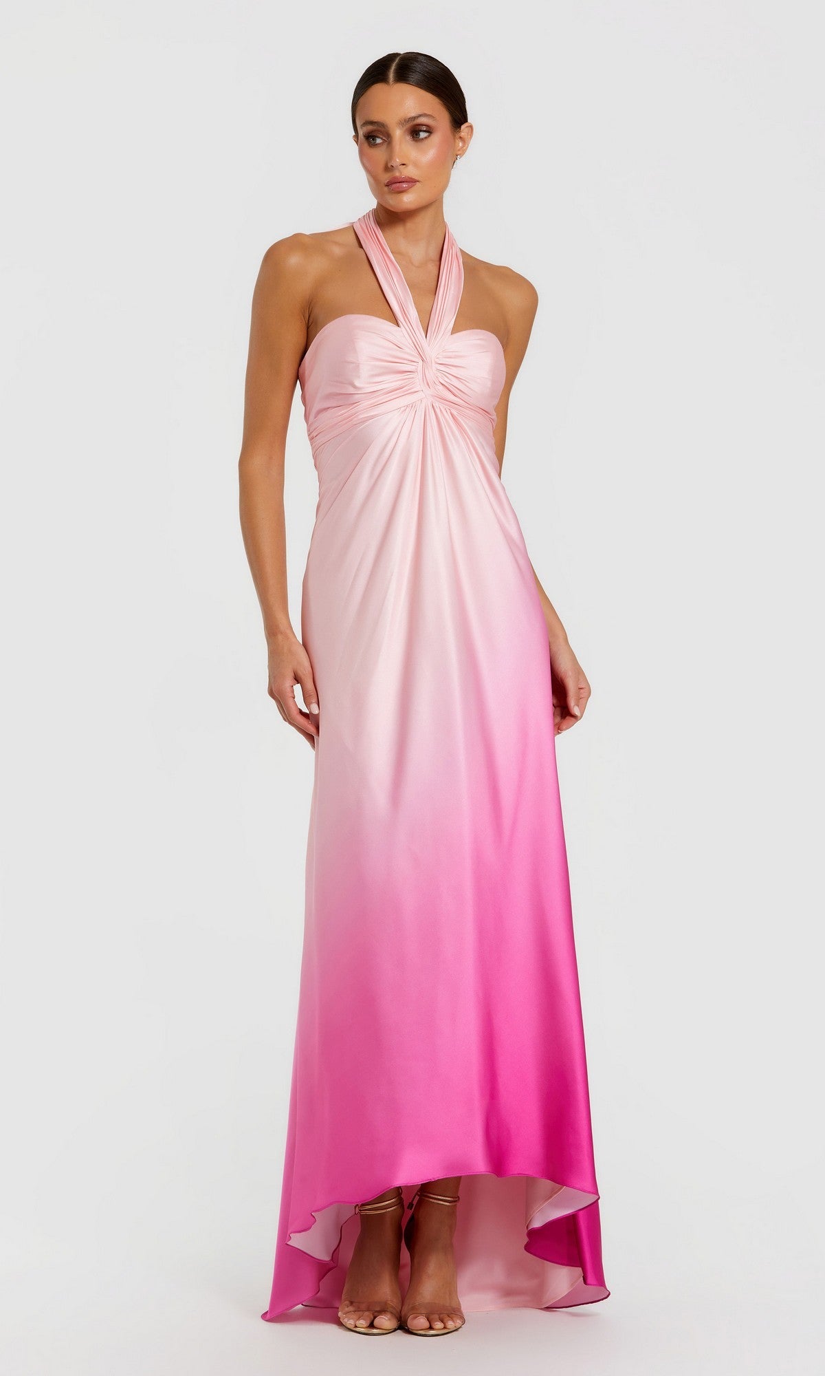 A woman stands against a white background wearing the Mac Duggal 68501—a sleeveless, halter-neck, floor-length formal dress in pink ombre charmeuse with a gathered bodice.